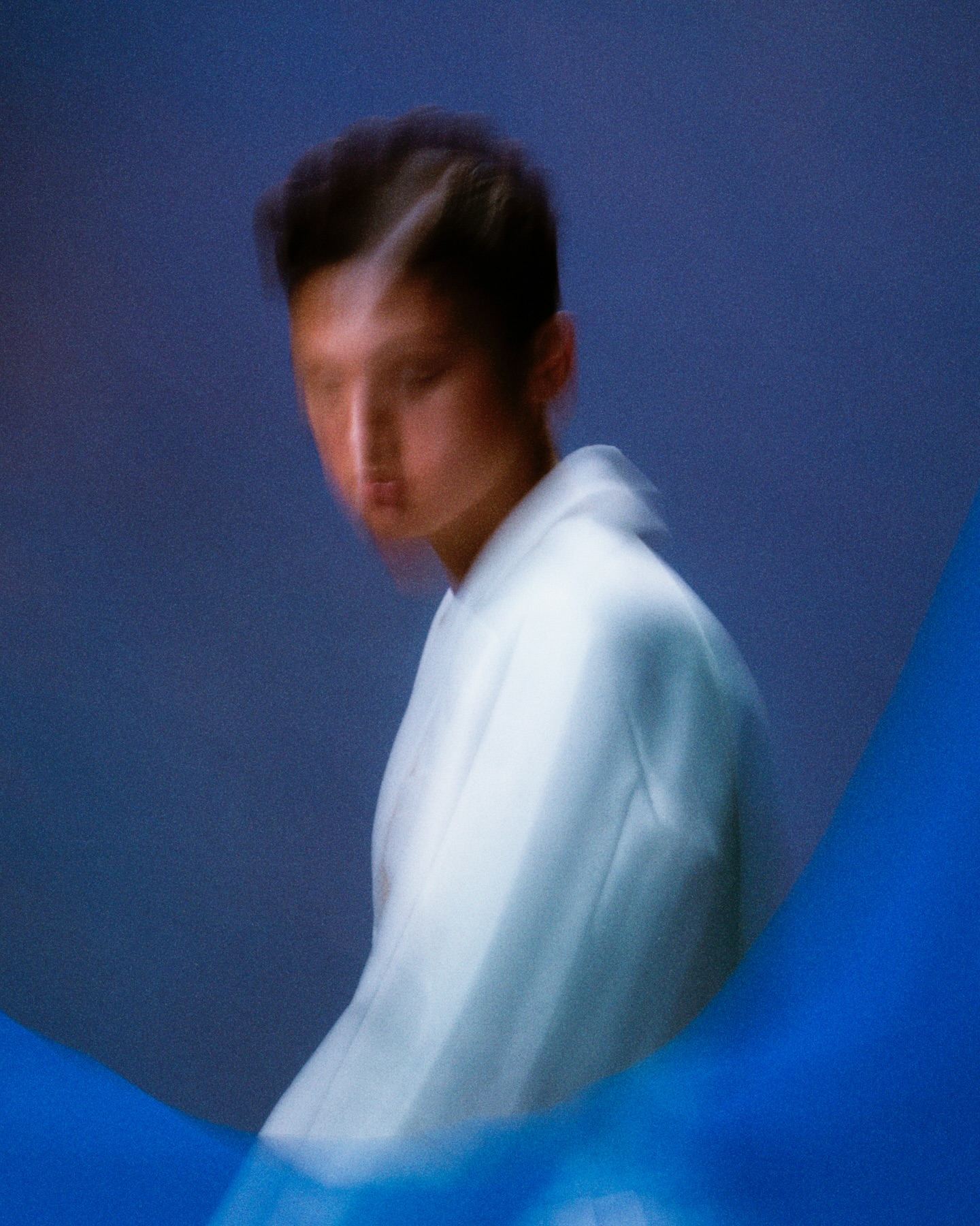 Portrait of an East Asian person in a white jacket against a blue backdrop. Motion blur adds an ethereal effect to the image.