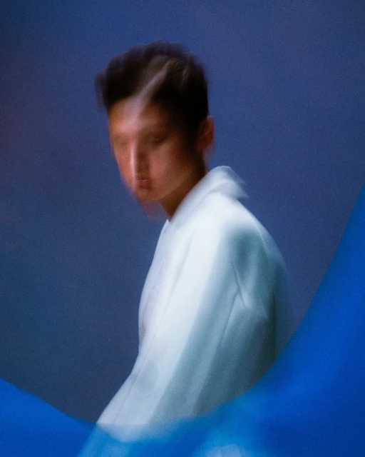 Portrait of an East Asian person in a white jacket against a blue backdrop. Motion blur adds an ethereal effect to the image.