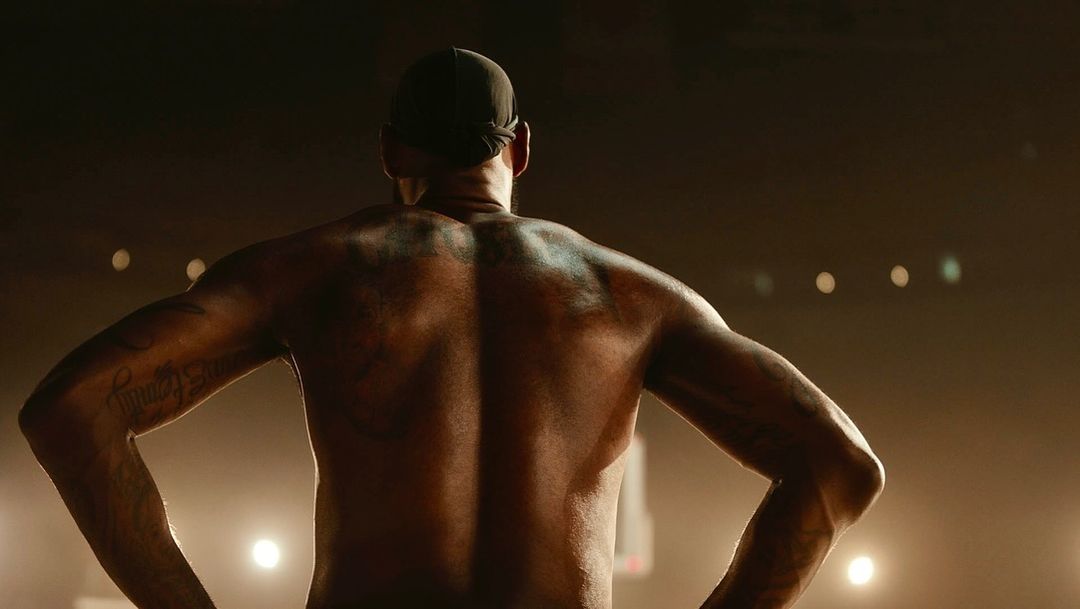 Athletic, muscular adult male in a cap stands with hands on hips, showcasing a heavily tattooed back in a backlit stadium setting.