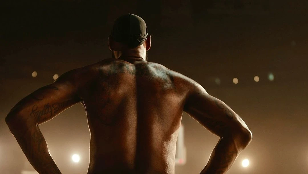 Athletic, muscular adult male in a cap stands with hands on hips, showcasing a heavily tattooed back in a backlit stadium setting.
