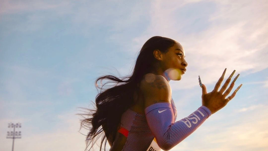 Track athlete running with long nails against a blue sky. The runner's arm sleeves feature the Nike swoosh and the letters USA.