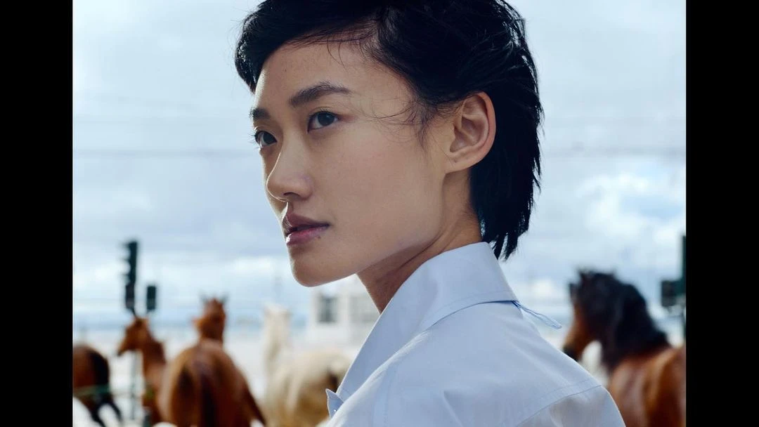 Portrait of an Asian woman in a white shirt with horses in the background under a cloudy sky; shot in natural light.