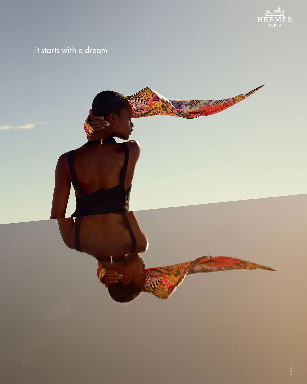 Fashion shot of a dark-skinned woman with a colorful scarf blowing in the wind, mirrored in a reflective surface against a blue sky.