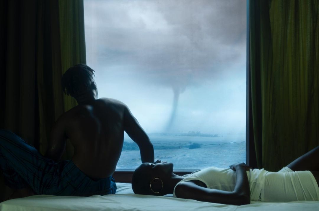 Intimate portrait of a couple, one seated and one reclining on a bed, silhouetted against a window revealing a distant tornado.