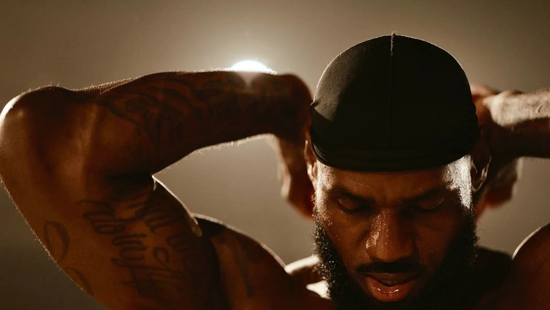 Close-up of muscular adult male athlete with tattoos wearing a black head covering, with arms raised behind his head in a neutral indoor setting.