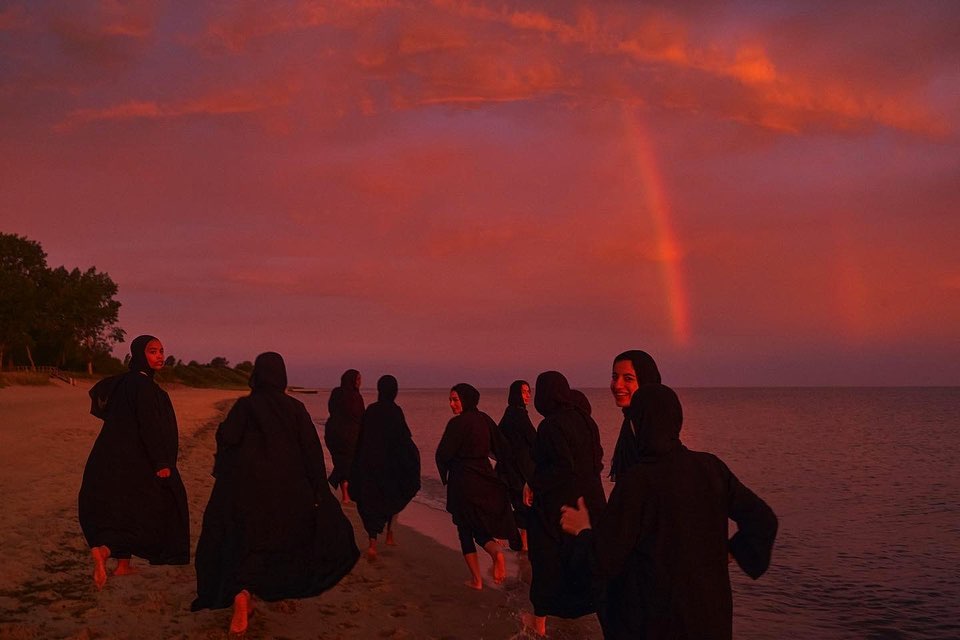 Group of women in black abayas walking along a sandy beach under a dramatic reddish sunset sky with a faint rainbow