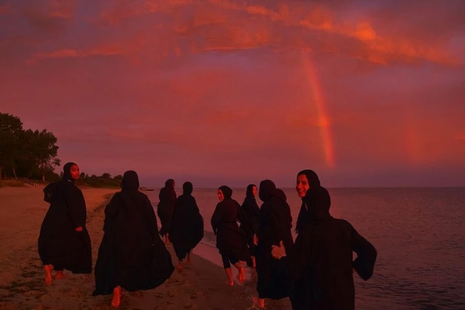 Group of women in black abayas walking along a sandy beach under a dramatic reddish sunset sky with a faint rainbow