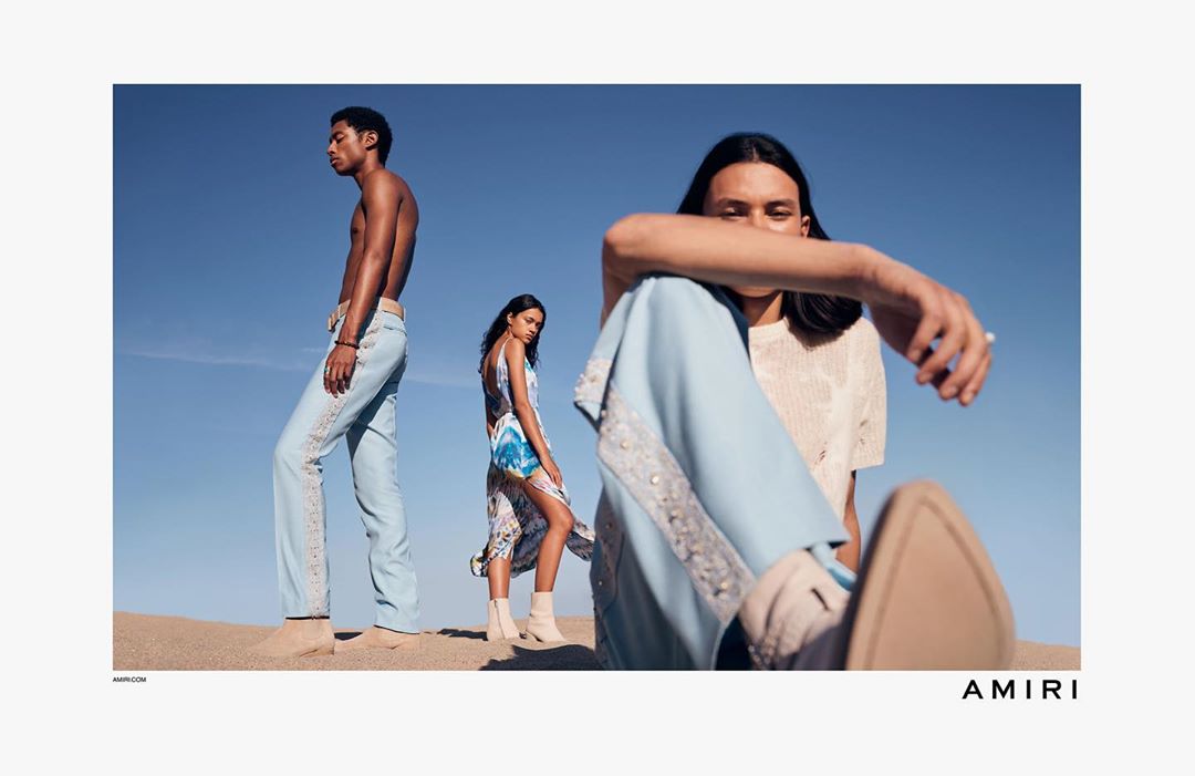 Fashion shot featuring three models on a sandy dune under a clear blue sky, showcasing light-colored, patterned clothing and stylish, desert-chic attire.