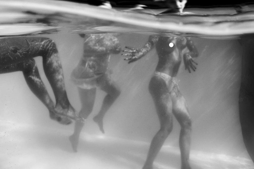 Monochrome underwater photograph of three figures submerged in a pool, creating an ethereal and slightly surreal black and white image.