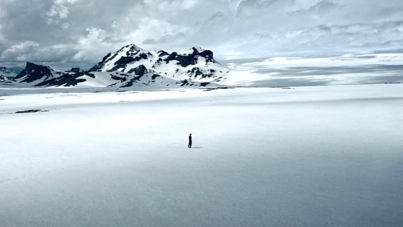 A lone small figure standing on a vast snowy expanse beneath a dramatic mountain range under an overcast sky.