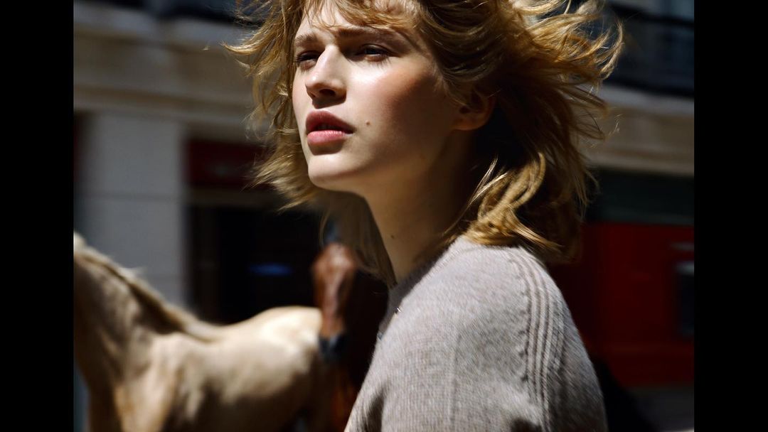 Outdoor portrait of a young fair-skinned woman with windswept blond hair, wearing a grey sweater, with a horse in the background.