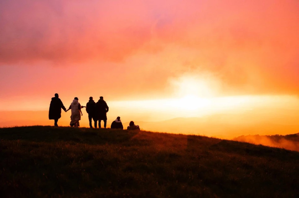 Group of silhouetted people stands on a grassy hill against a vibrant orange and pink sunset sky with some clouds and distant hills.