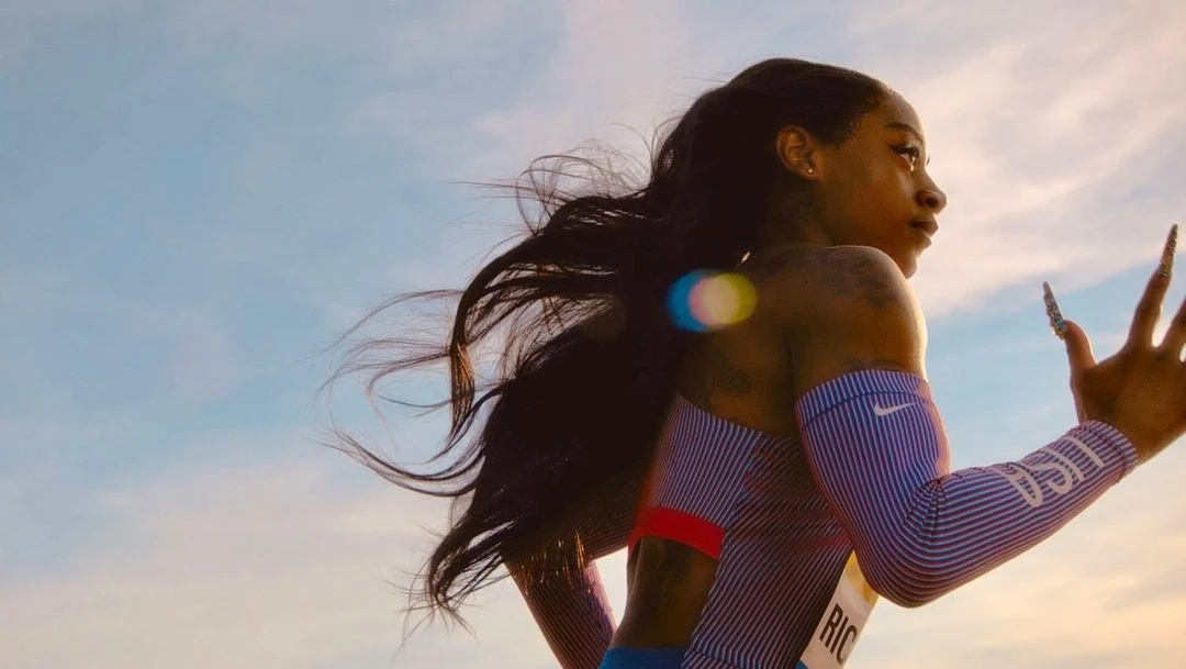 Portrait of track athlete Sha'Carri Richardson against blue sky, wearing blue and white striped athletic gear, wind blowing her hair.