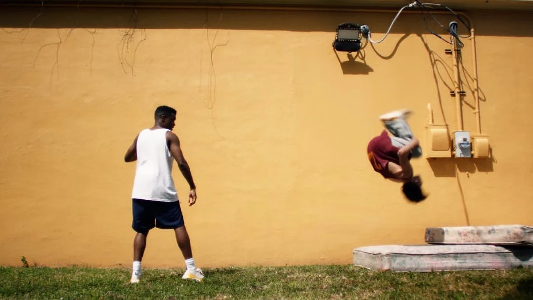 Two people outside. One person stands on the ground. The other person flips in the air, with stacked mattresses nearby.