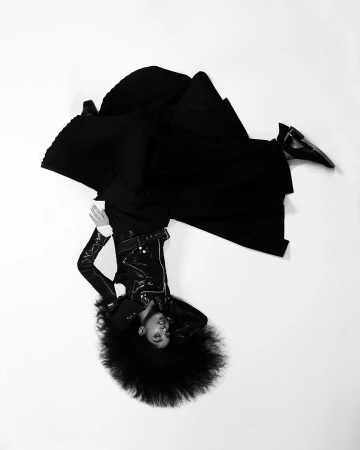 Model with voluminous dark hair wearing a black jacket and voluminous skirt lying on a white background.