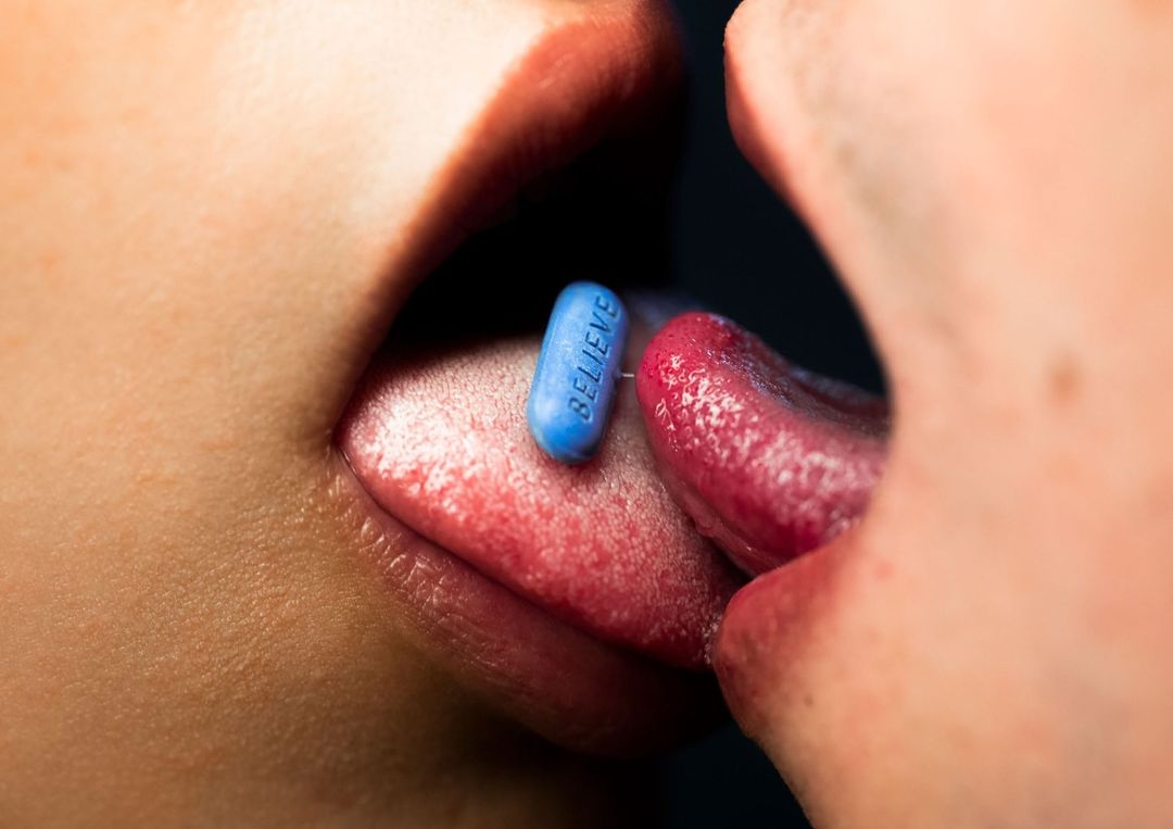 Macro shot of two people's lips and tongues touching, with a blue pill that says 'Believe' on one tongue in a studio with dark backdrop.