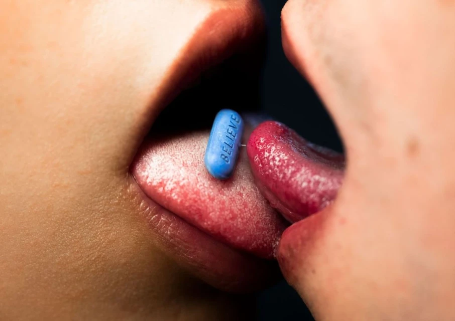 Macro shot of two people's lips and tongues touching, with a blue pill that says 'Believe' on one tongue in a studio with dark backdrop.