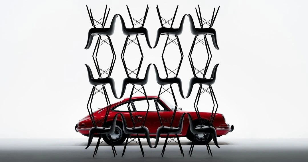 A red classic Porsche sits between a wall of black Eames chairs stacked and arranged in an evenly spaced geometric pattern.