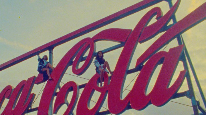 Two women are posing on a large red Coca-Cola sign outdoors under a sky with high horizon. The sign is the primary focus of the image.