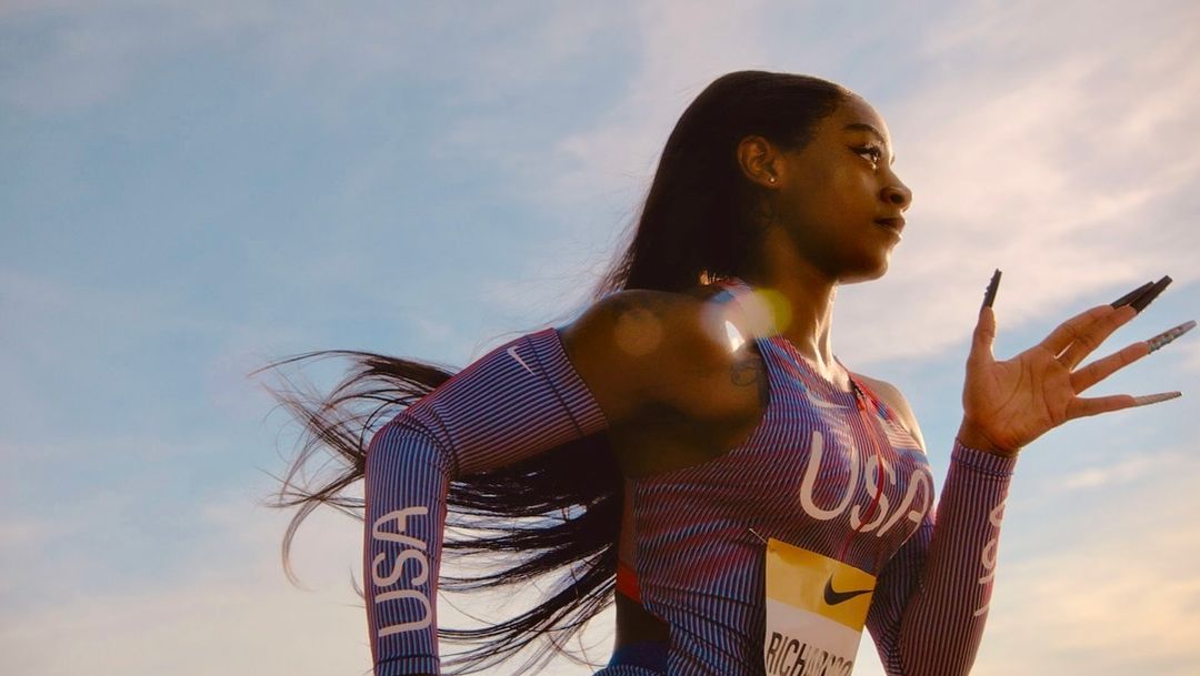 Athlete Sha'Carri Richardson in uniform with 'USA' lettering and long nails stands against a sunny sky in a medium portrait shot.