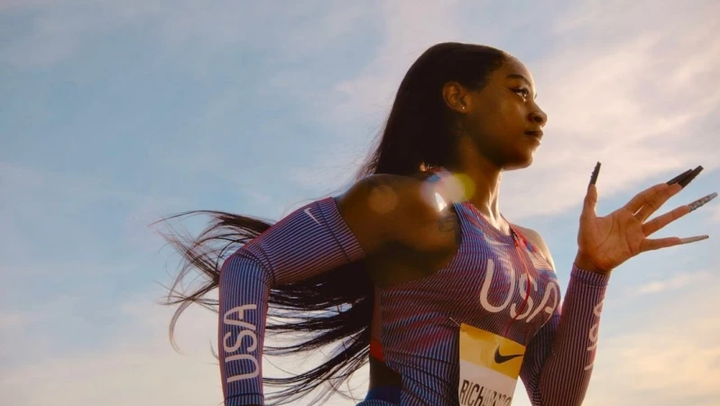 Athlete Sha'Carri Richardson in uniform with 'USA' lettering and long nails stands against a sunny sky in a medium portrait shot.