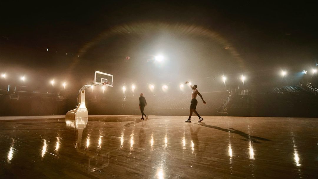 Two athletes on a brightly lit basketball court with stadium seating, one shirtless and throwing the ball, the other walking.
