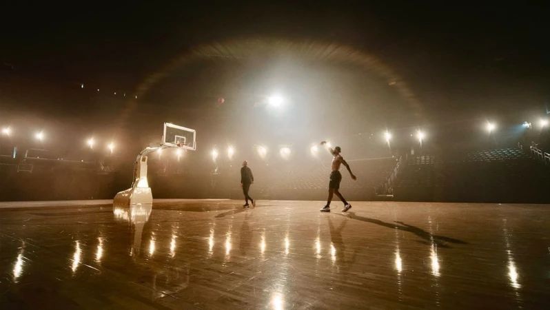 Two athletes on a brightly lit basketball court with stadium seating, one shirtless and throwing the ball, the other walking.