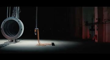 Abstract industrial scene featuring a metal ring suspended by cables above a drain with liquid, set in a dimly lit, cavernous interior.