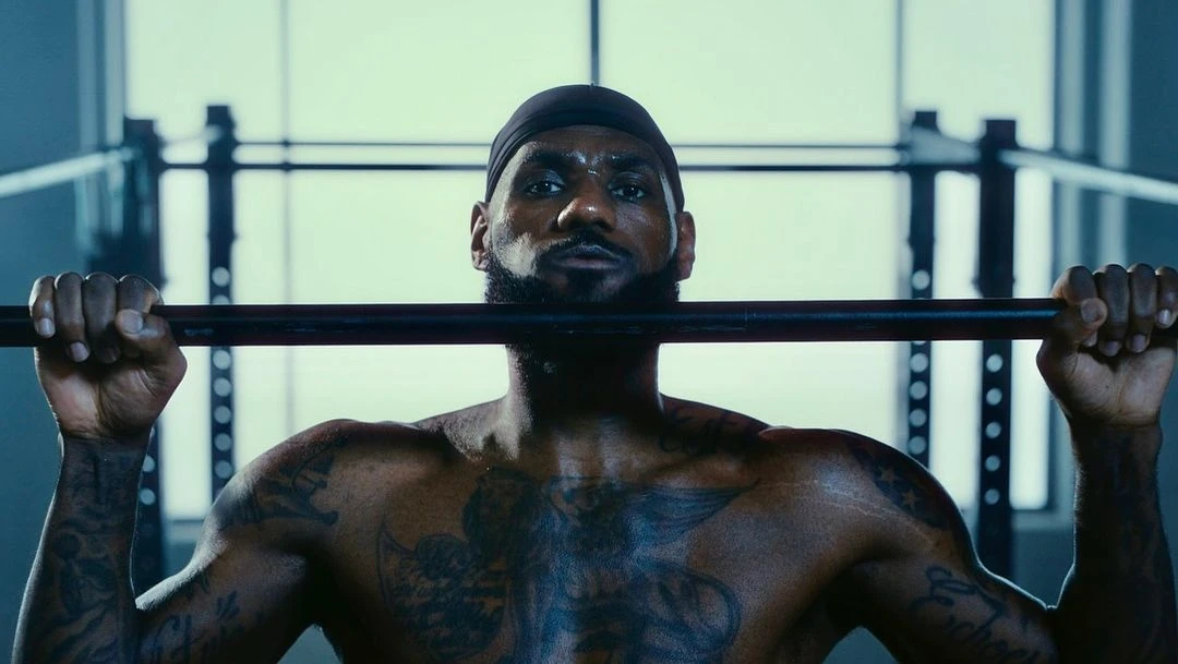 Shirtless adult male with dark hair and facial hair, wearing a black bandana, gripping a black metal bar in a fitness setting.
