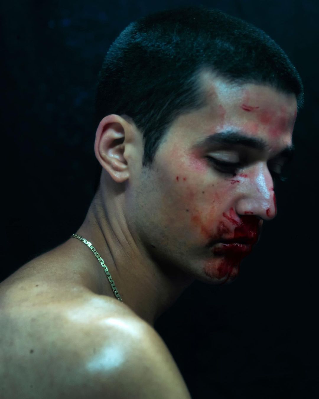 Close-up shot of a young adult man with blood on his face and neck, wearing a gold chain, against a dark, indistinct background.
