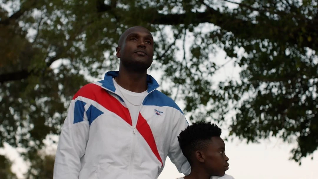 Adult male and child pose outdoors in front of leafy green trees. The man wears a white Reebok jacket with blue and red accents.