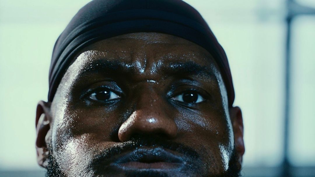 Close-up portrait of a sweaty, dark-skinned adult male athlete wearing a black head covering in a gym setting with natural lighting.