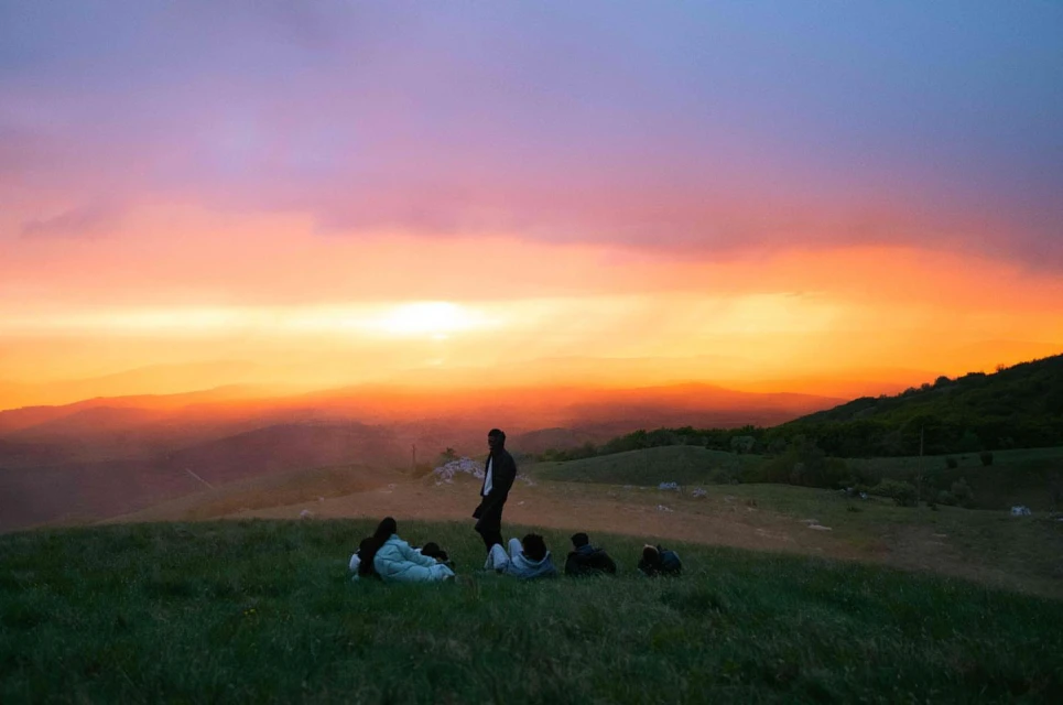 Group of friends relax on a grassy hill at sunset with mountains in the distance under a vibrant, colorful sky.