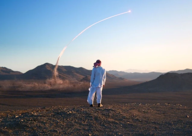 Man in traditional Saudi clothing stands in desert landscape, watching a missile launch with a vapor trail against a blue sky.