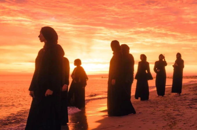 Group of people in dark clothing walk on a sandy beach during a vibrant sunset, silhouetted against the colorful sky and calm ocean.