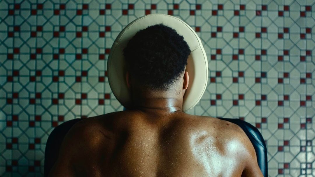 Back view of a shirtless adult male seated in a chair with a headrest, set against a backdrop of tiled walls with a geometric pattern.