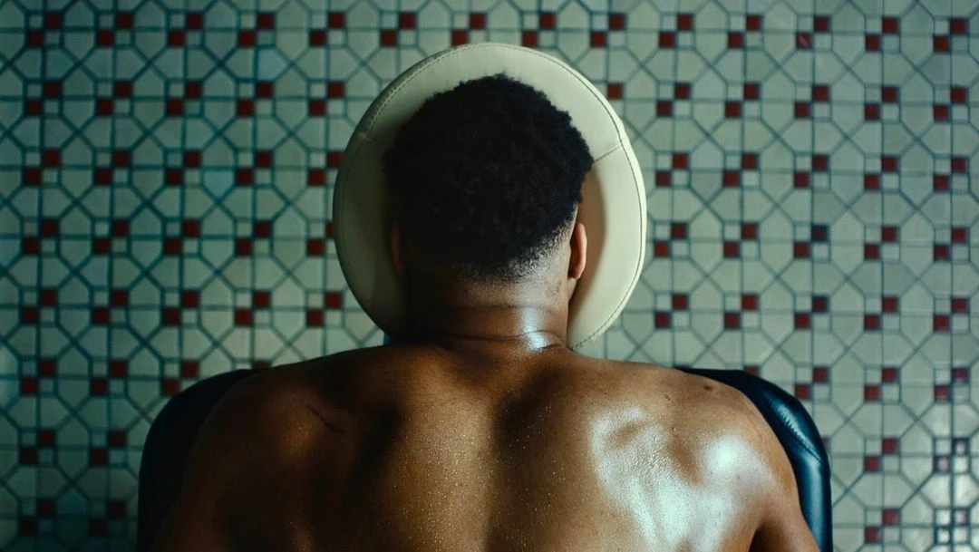 Back view of a shirtless adult male seated in a chair with a headrest, set against a backdrop of tiled walls with a geometric pattern.