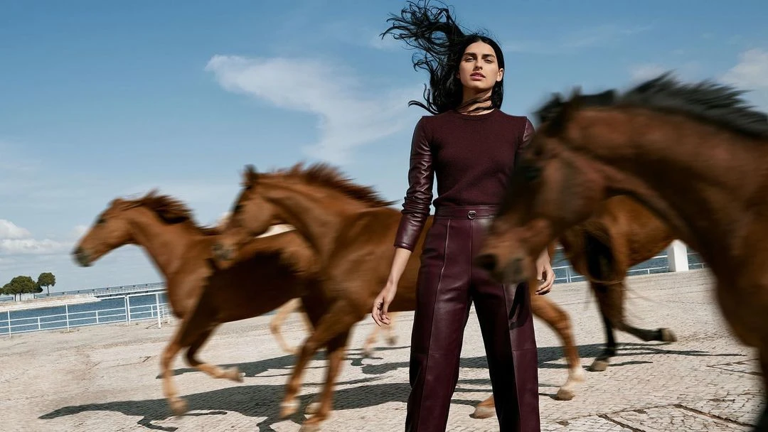 Woman in burgundy outfit poses in front of running horses in an outdoor fashion editorial on a paved surface under a blue sky.