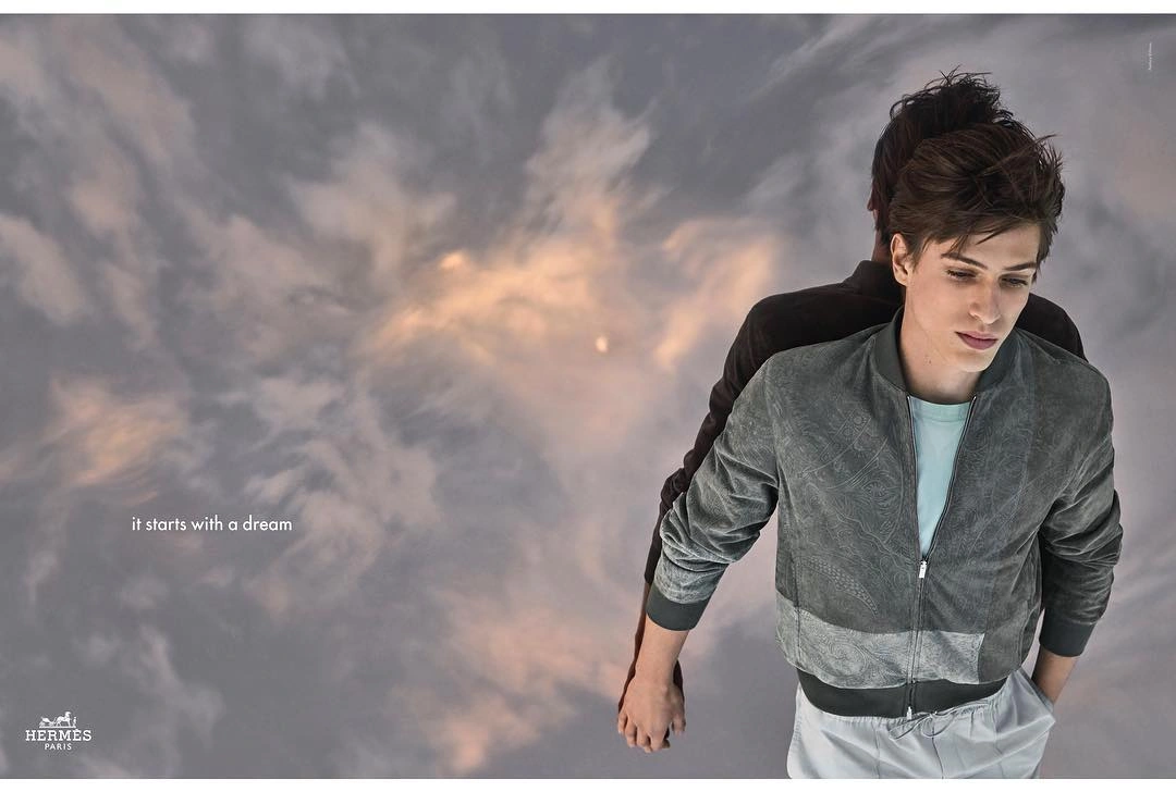 Two young men holding hands against a swirling cloudy sky background, wearing grey jackets.