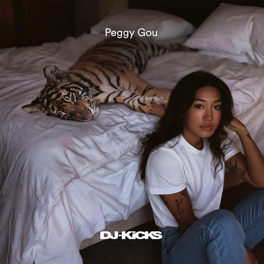 Woman in white t-shirt and blue jeans sitting on the floor next to a bed with a resting tiger