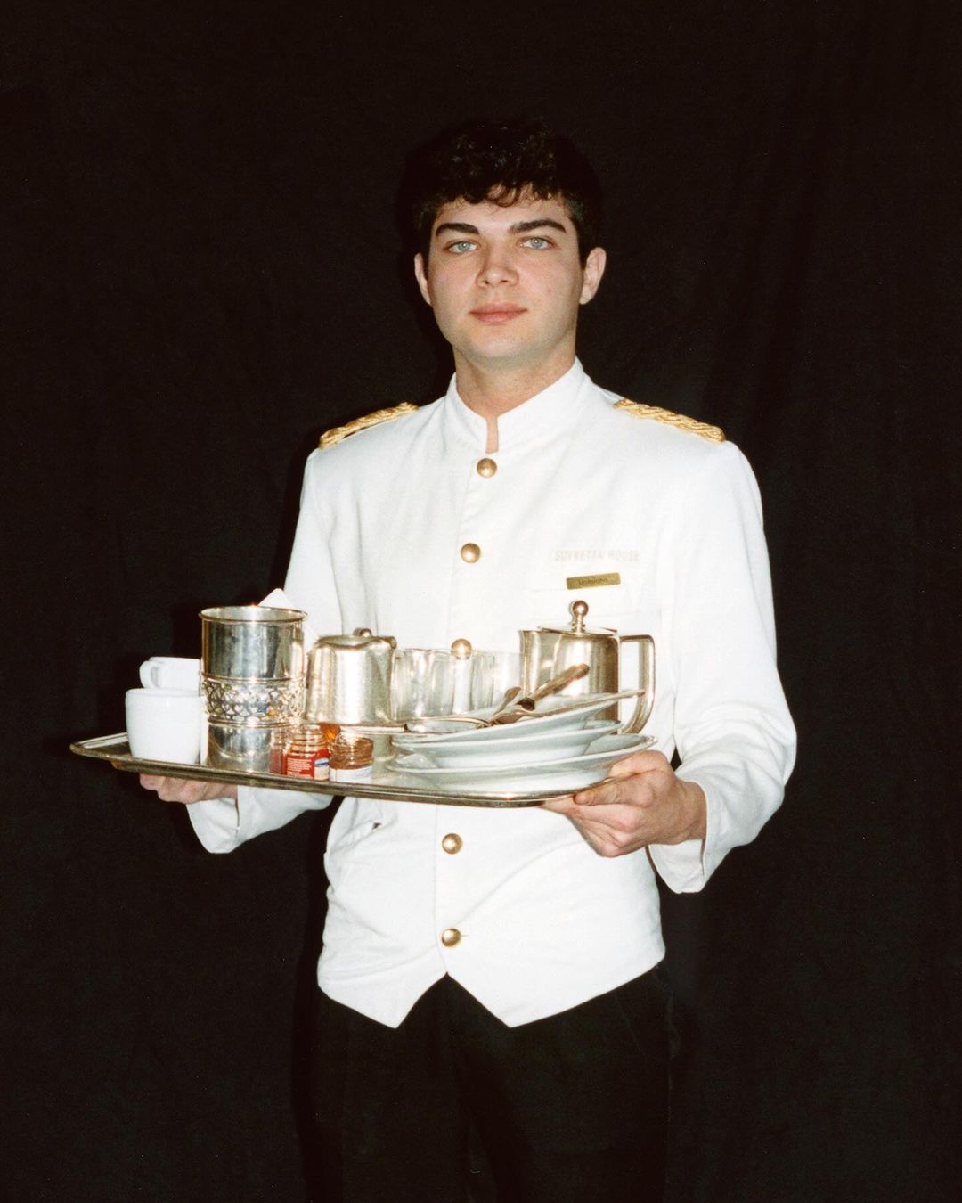 Server holding tray of tableware in a vintage style. White uniform with black pants against black background. Indoor, studio.
