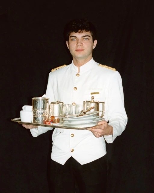 Server holding tray of tableware in a vintage style. White uniform with black pants against black background. Indoor, studio.