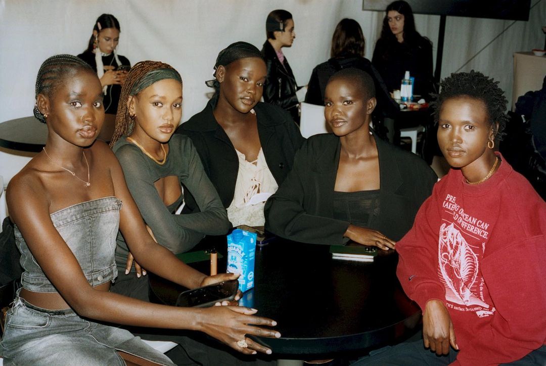 Group of African female fashion models gather around a dark table in an indoor setting, wearing stylish clothing and accessories, posing.