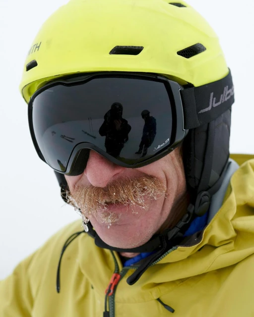 Close-up of a person with a large mustache wearing a yellow ski helmet and goggles with a reflection of skiers.
