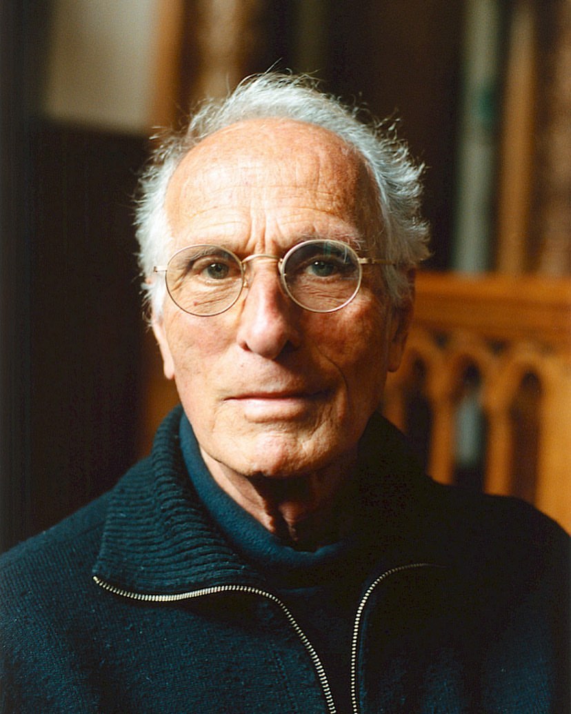 Portrait of an elderly Caucasian man with glasses wearing a dark sweater, set against a softly blurred, dark background and carved wood.