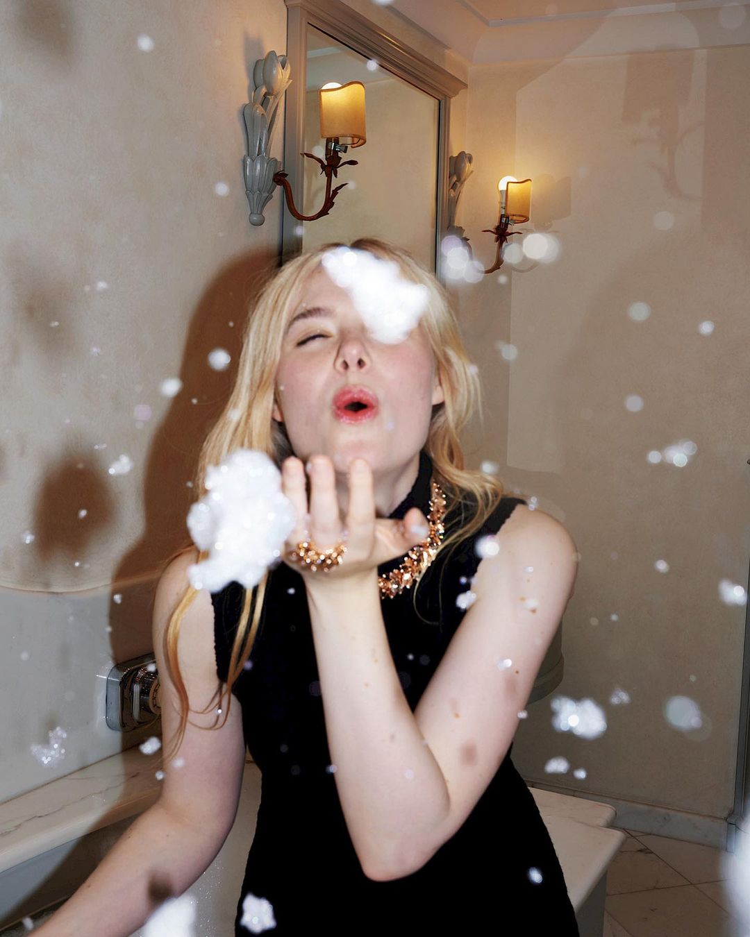 A young woman with blonde hair wearing a black dress and gold necklace blows foam from her hand in a bathroom-like indoor setting.