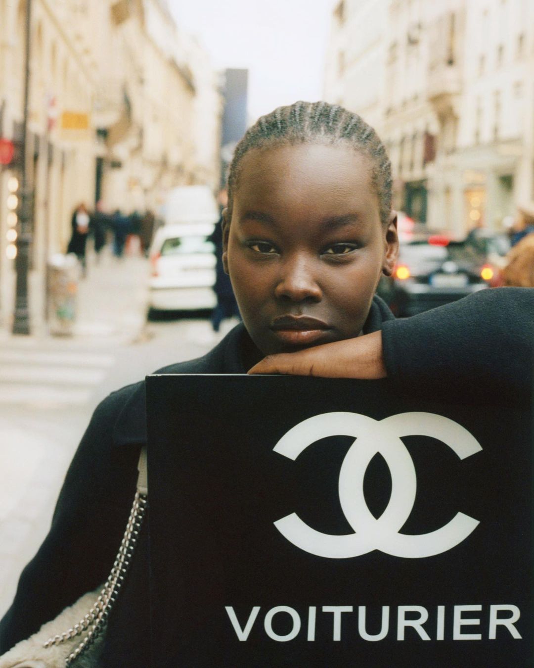 Portrait of a young African woman in a black coat holding a black sign with the white Chanel logo and the text VOITURIER on a city street.