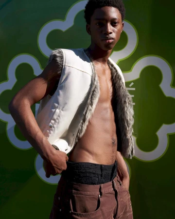 Young black man wearing a white faux-fur vest and brown trousers posing outdoors in Paris by photographer Jonas Unger.