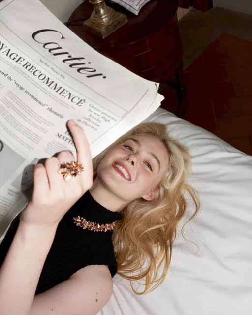 Elle Fanning poses with Cartier jewelry and newspaper, lying on a white bed with a vintage bedside table, bathed in soft, natural light.