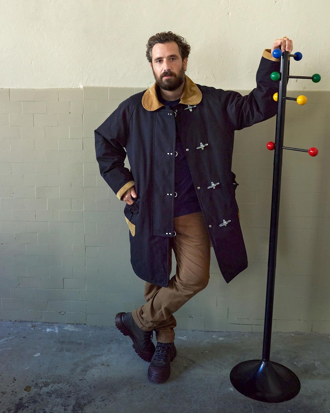 Adult man with a beard wearing a black toggle coat leans against a colorful coat rack inside a room with a concrete floor and wall.